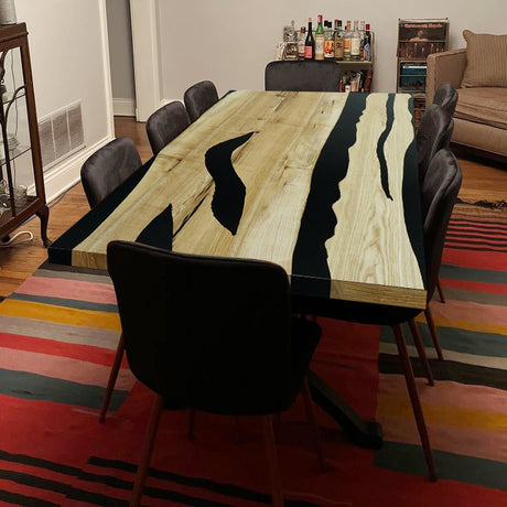Rectangular dining table with light - toned wood and black epoxy - resin inlays, surrounded by black chairs on a colorful rug.