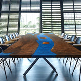 Rectangular conference table with a wooden surface and blue epoxy - resin inlay, surrounded by black chairs in a room with large windows.