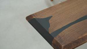 A long straight-edge dining or coffee table made of wood with black resin inlays, accompanied by a uniquely designed wooden chair with resin details