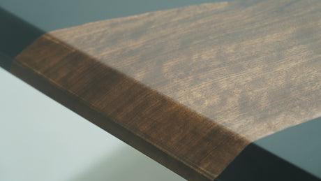 A dining table made of walnut wood with dark black resin inlays, supported by clear acrylic legs, and part of a wooden chair visible in the background